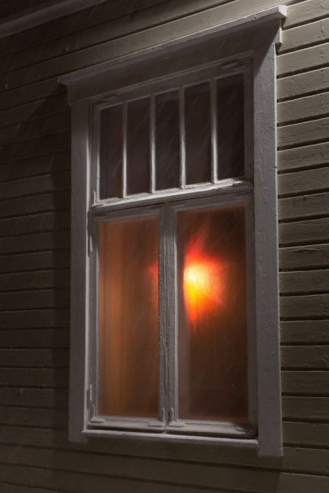 Vanhan puutalon ikkuna pimeästä ulkoapäin katsottuna. Sisällä palaa valo