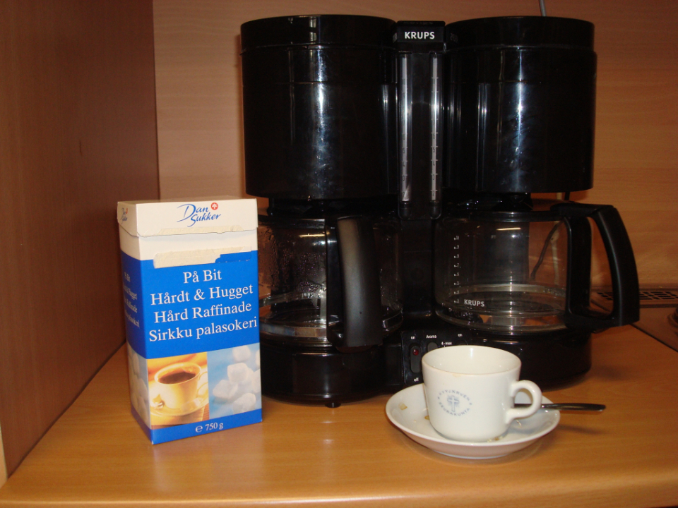 Kuvassa kahvikuppi, kahvin keitin ja sokeripaketti
